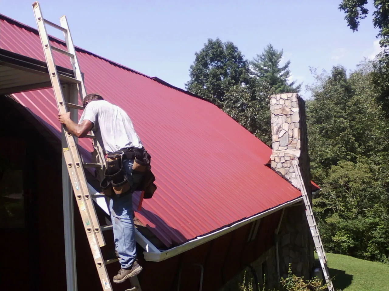 Roofer on a ladder beside a red metal roof and stone chimney — Soffit & Fascia Repair by Roofing Brothers in Limestone